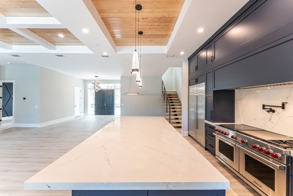 Modern kitchen in homes in Nocatee FL with a large marble island, stainless steel appliances, dark cabinetry, pendant lights, wooden ceiling accents, and open space leading to stairs and the front door.