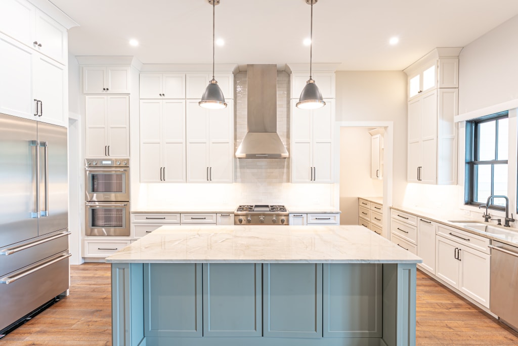 Bright, modern kitchen in one of the stunning New Homes in Ponte Vedra Beach FL, featuring white cabinets, stainless steel appliances, a large marble island, pendant lights, wood flooring, and a window above the sink for natural light.