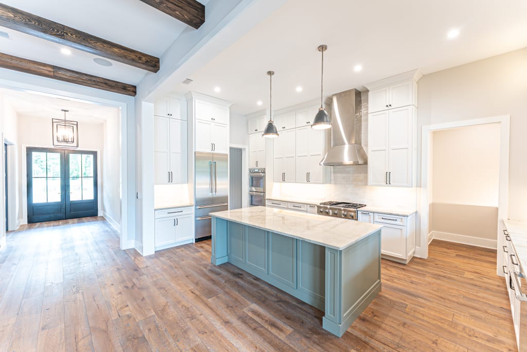 Bright modern kitchen in one of the stunning new homes in Ponte Vedra Beach FL, featuring white cabinets, stainless steel appliances, a marble-topped island with light blue base, wood floors, pendant lights, and exposed wooden ceiling beams.