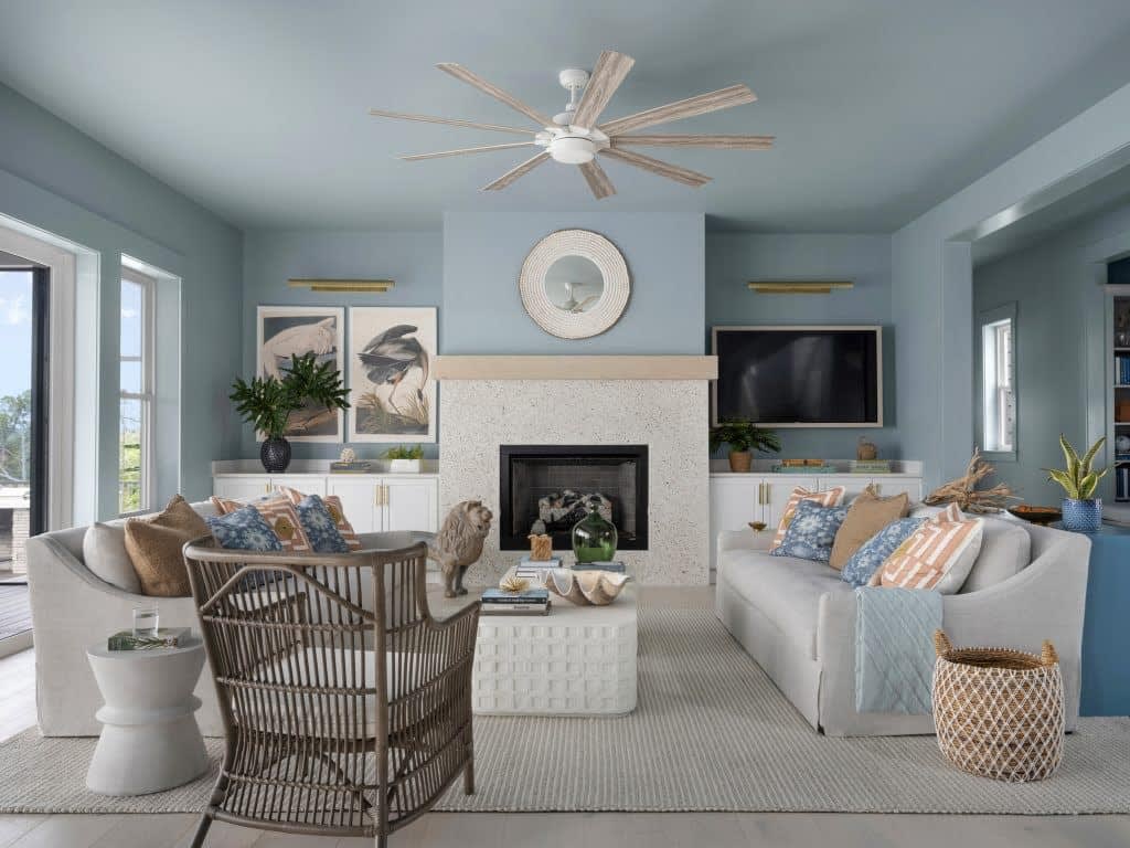 A stylish living room with light blue walls, a gray sofa, patterned cushions, wicker chairs, a fireplace, plants, a large ceiling fan, wall art, a TV, and large windows letting in natural light.