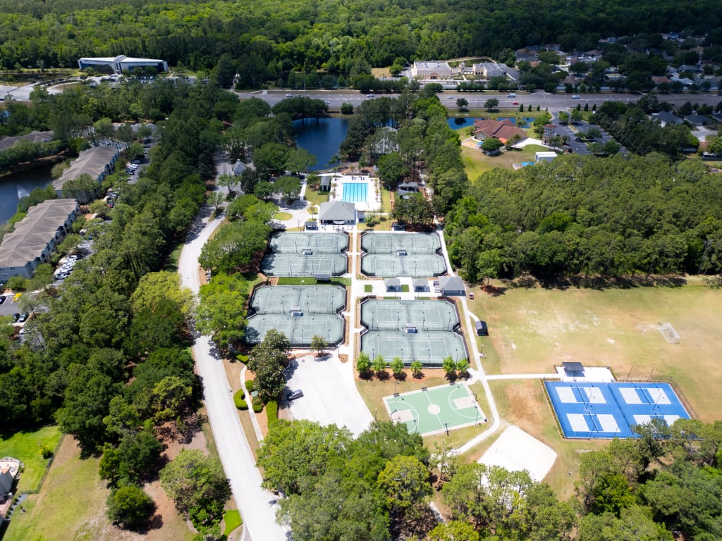 Aerial view of a sports complex with multiple tennis courts, basketball courts, and pickleball courts, surrounded by trees, a swimming pool, and nearby new homes in Ponte Vedra Beach FL.