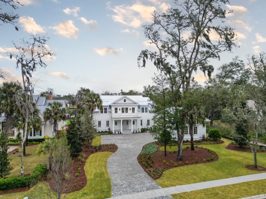 A large white two-story house with columns and a front porch, surrounded by trees and landscaped lawns—a stunning example of homes in Nocatee FL—with a curved stone driveway leading to the entrance under a partly cloudy sky.