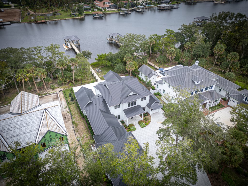 Aerial view of modern homes in Nocatee FL with gray roofs near a river, surrounded by trees and docks, and one house under construction on the left side.