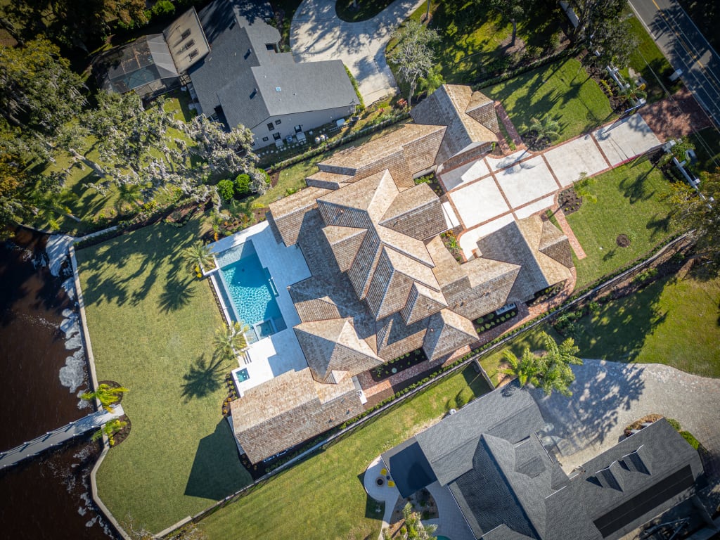 Aerial view of a large, luxurious house with a complex roof design, a swimming pool, spacious green lawn, long driveway, and surrounding trees, situated next to a body of water.