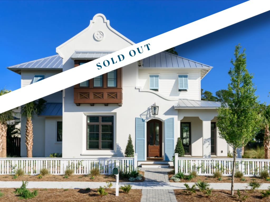 A modern two-story white house with a gated front yard and blue shutters, viewed on a sunny day—perfect for those seeking homes in Jacksonville Beach FL. A white diagonal banner across the image reads SOLD OUT in bold blue letters.