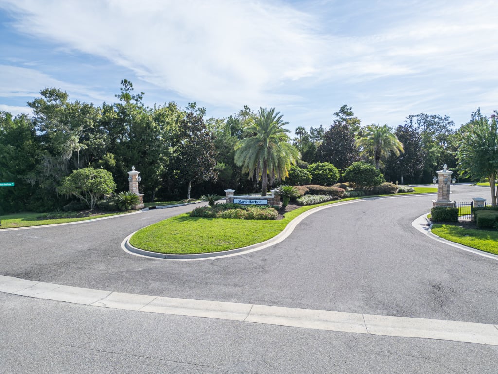 A circular driveway with a sign in the center, surrounded by green grass, palm trees, and shrubs. Tall stone pillars mark the entrance, and trees line the background under a partly cloudy sky.