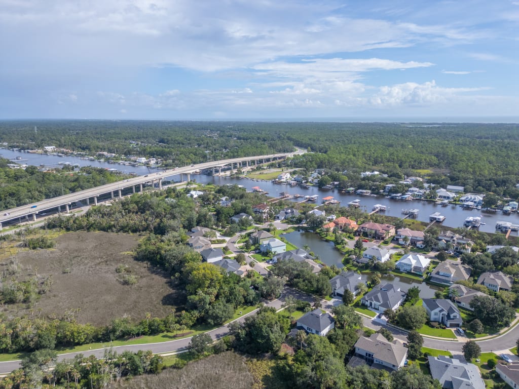Aerial view of a suburban neighborhood with a river, a bridge, homes with docks, green trees, and a partly cloudy sky in the background.