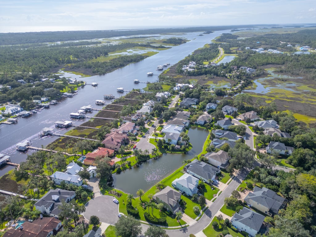 Aerial view of a suburban neighborhood with houses, green lawns, a pond, and a river lined with docks and boats, surrounded by trees and wetlands under a clear sky.