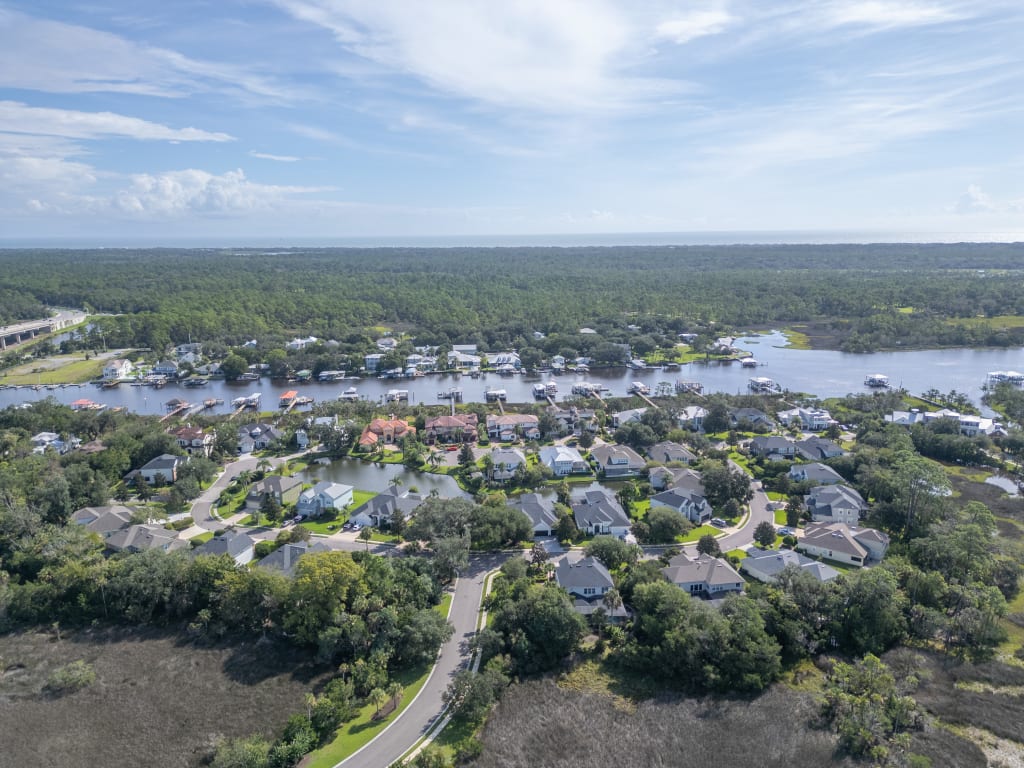 Aerial view of a suburban neighborhood with many houses, trees, and winding roads near a river or waterfront, with forested land and a partly cloudy sky in the background.