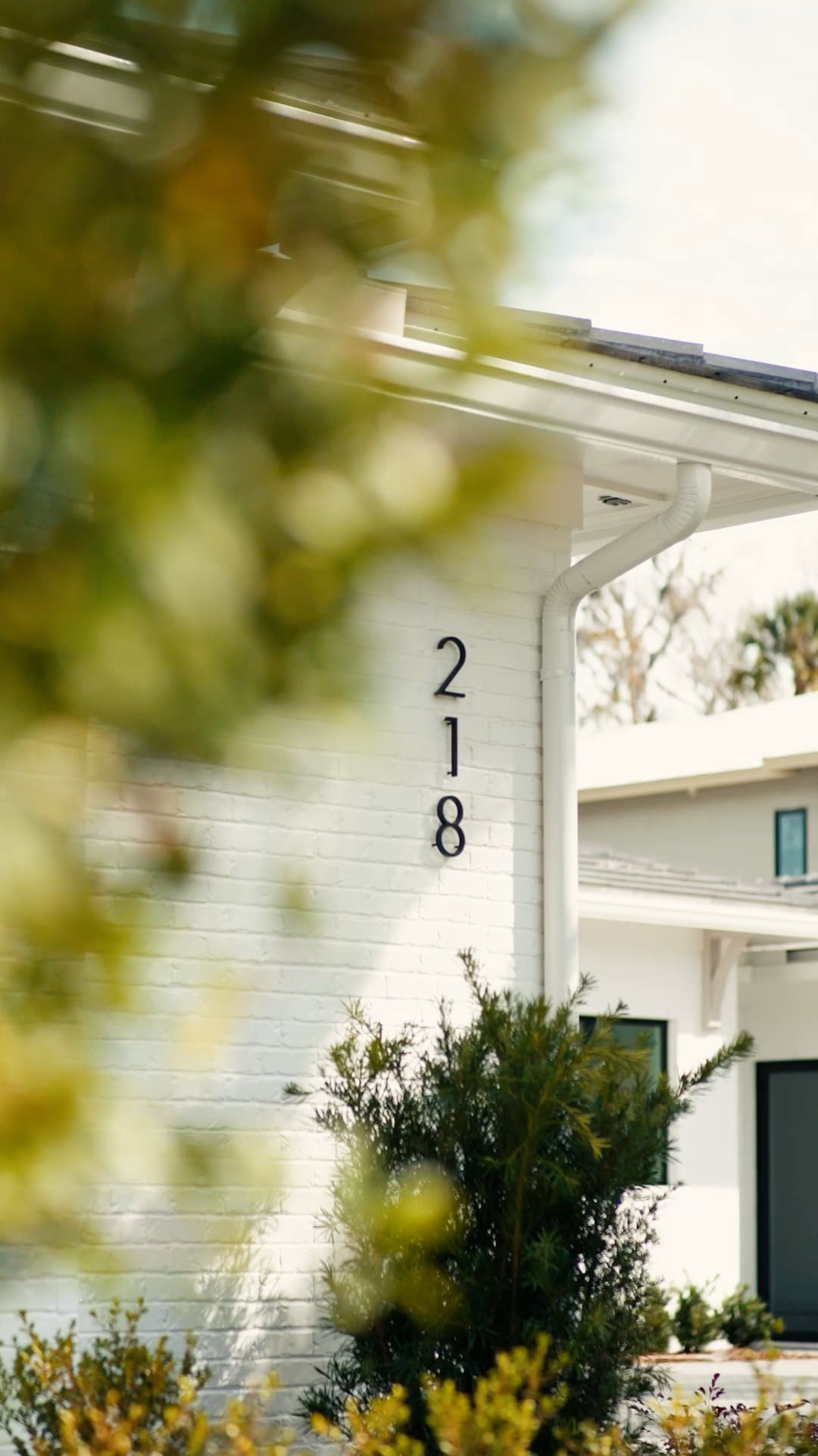 A white brick house with the number 218 displayed vertically near the entrance, partially obscured by green foliage in the foreground. Showcasing the work of a custom home builder in Florida, another modern building is visible in the blurred background.