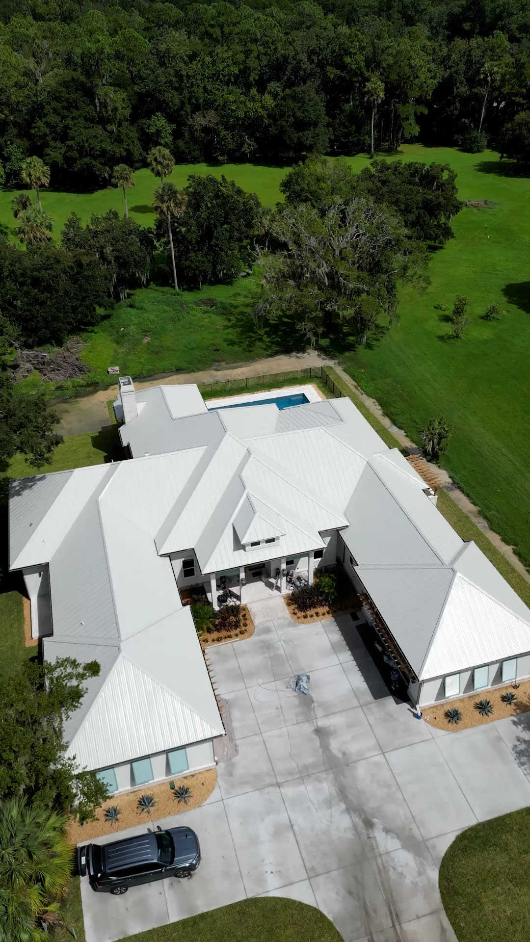 Aerial view of a large modern house by a custom home builder in Florida, featuring a white metal roof, multiple wings, spacious driveway, and backyard swimming pool, all nestled amid lush green grass and trees.