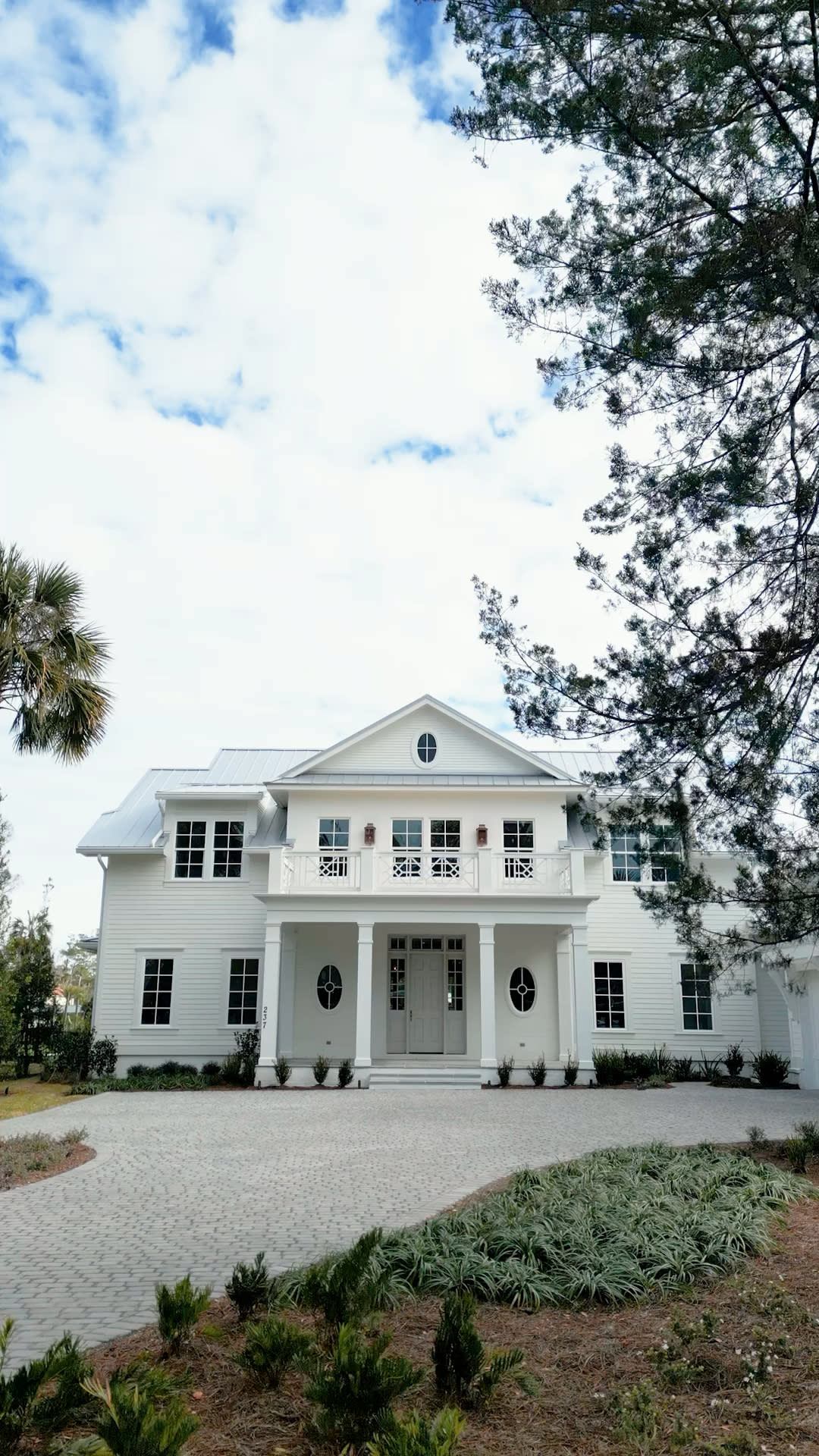 A large, two-story white house with a front porch and balcony, surrounded by trees and landscaping under a partly cloudy sky—crafted by a custom home builder in Florida. A paved driveway leads up to the entrance.