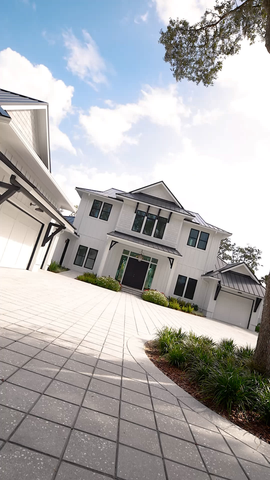 Modern two-story white house with black trim, large windows, and a spacious stone-paved driveway. Beautiful landscaping with green shrubs and trees showcases the detail you expect from a custom home builder in Florida. The sky above is mostly clear with some clouds.