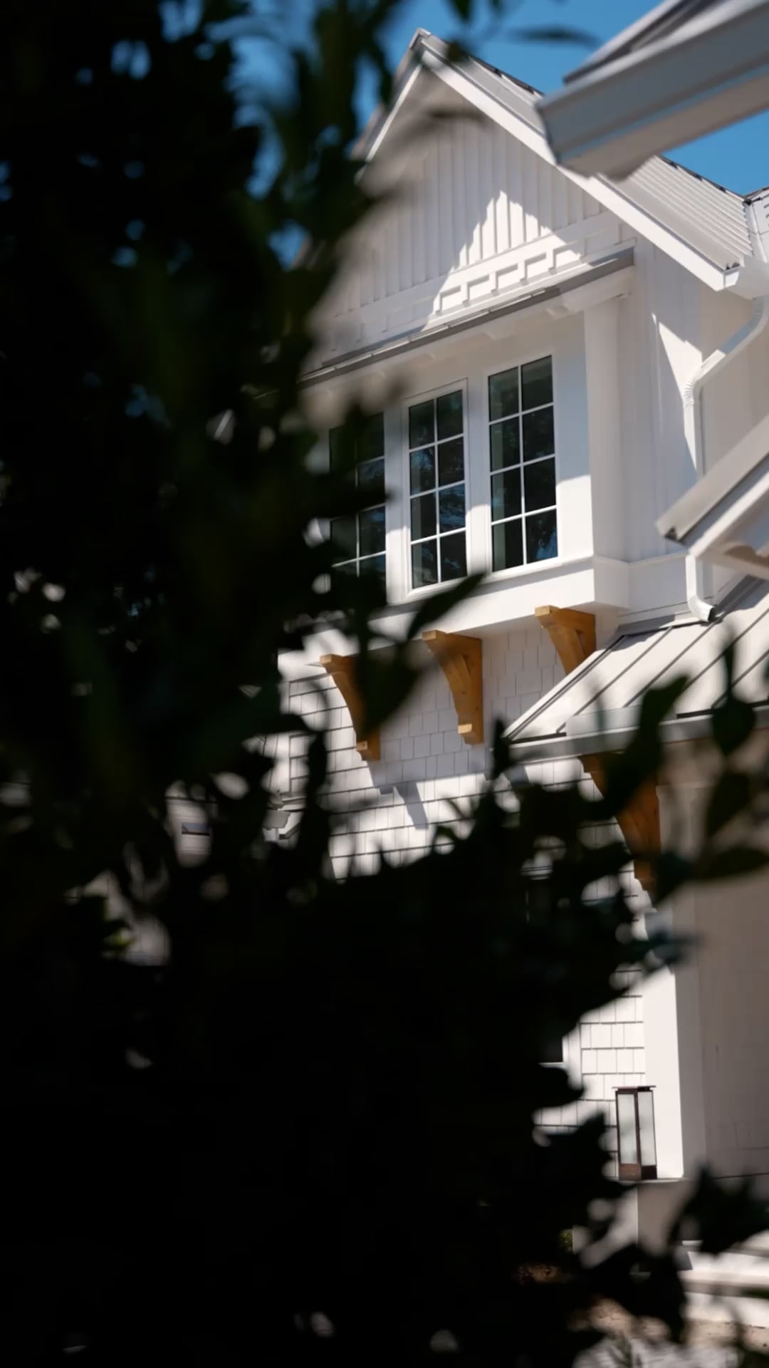 Partial view of a white house with large windows and wooden brackets, crafted by a custom home builder in Florida. Dark, leafy branches frame the home while sunlight highlights its upper facade.
