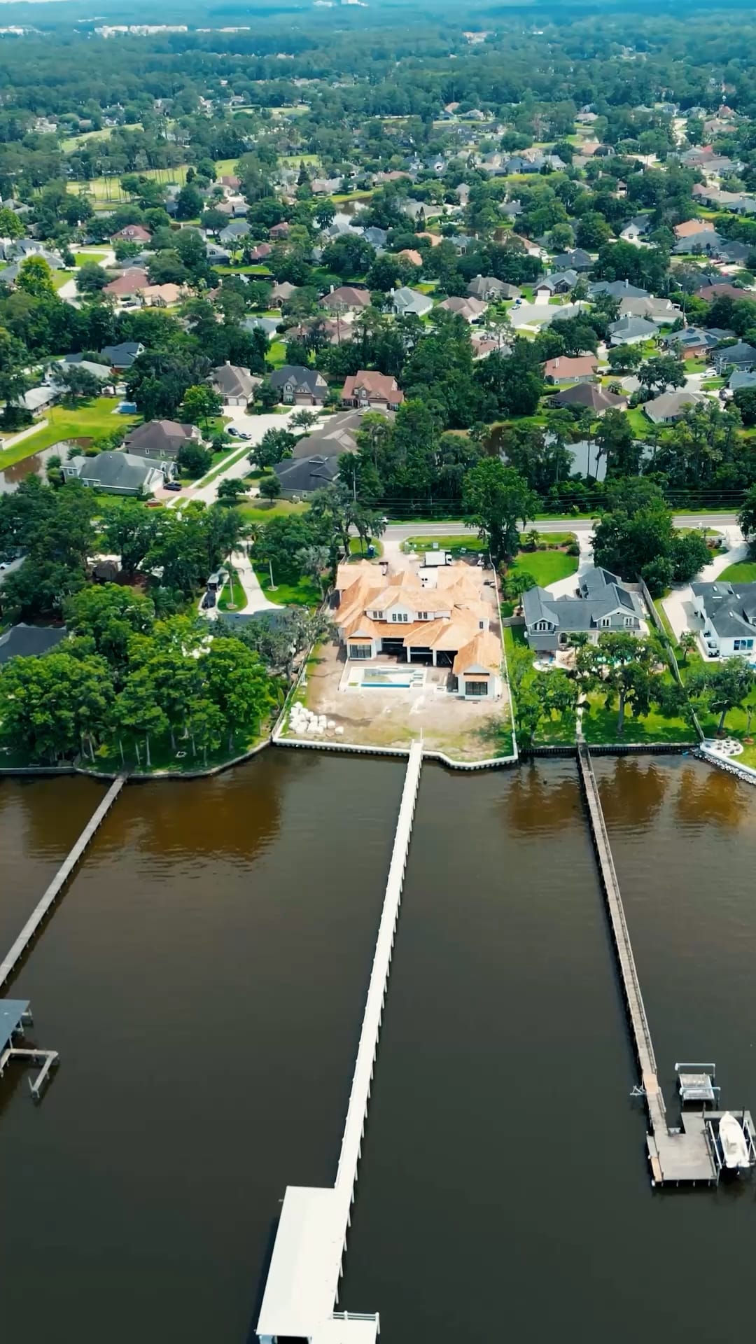 Aerial view of a suburban neighborhood with houses surrounded by trees, highlighting a large custom home builder in Florida project by the water, featuring a long private dock extending into a calm river with other docks along the shoreline.