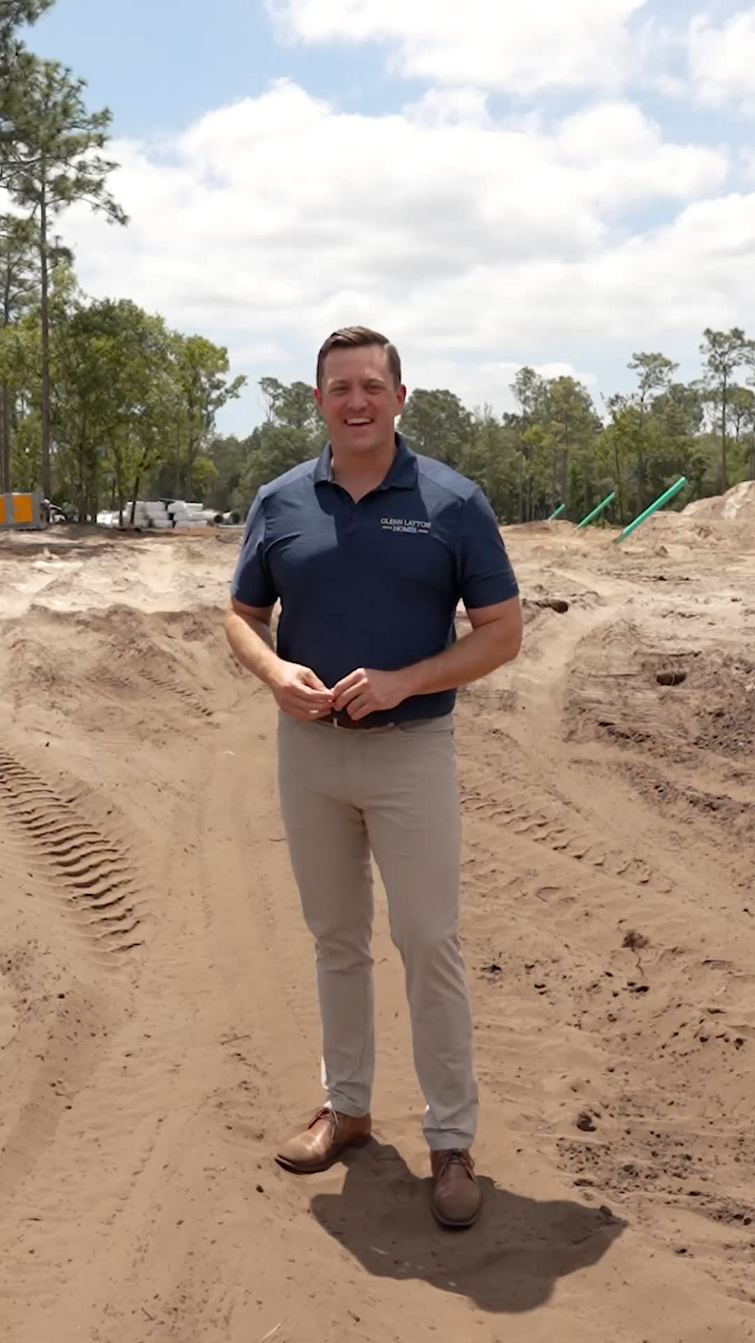 A man wearing a navy polo shirt and beige pants stands on a sandy construction site with tire tracks, surrounded by trees and cloudy blue sky—a typical scene for a custom home builder in Florida.