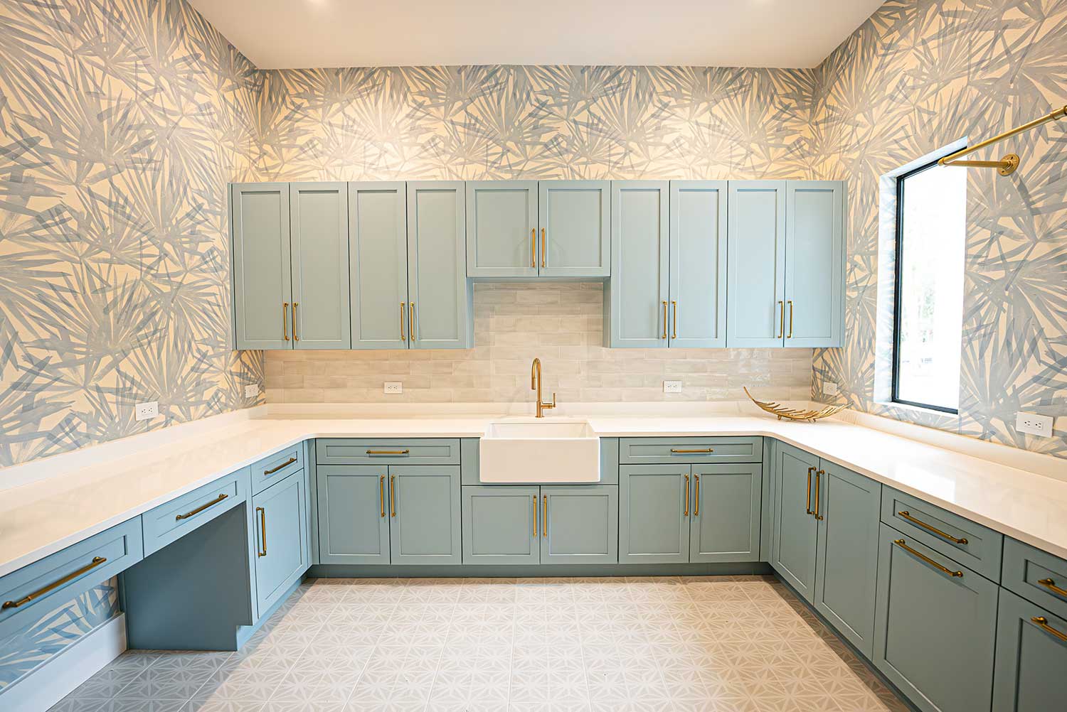 A modern kitchen with light blue cabinets, gold hardware, a large farmhouse sink, white countertops, patterned wallpaper, a tiled backsplash, a large window, and geometric tiled flooring.