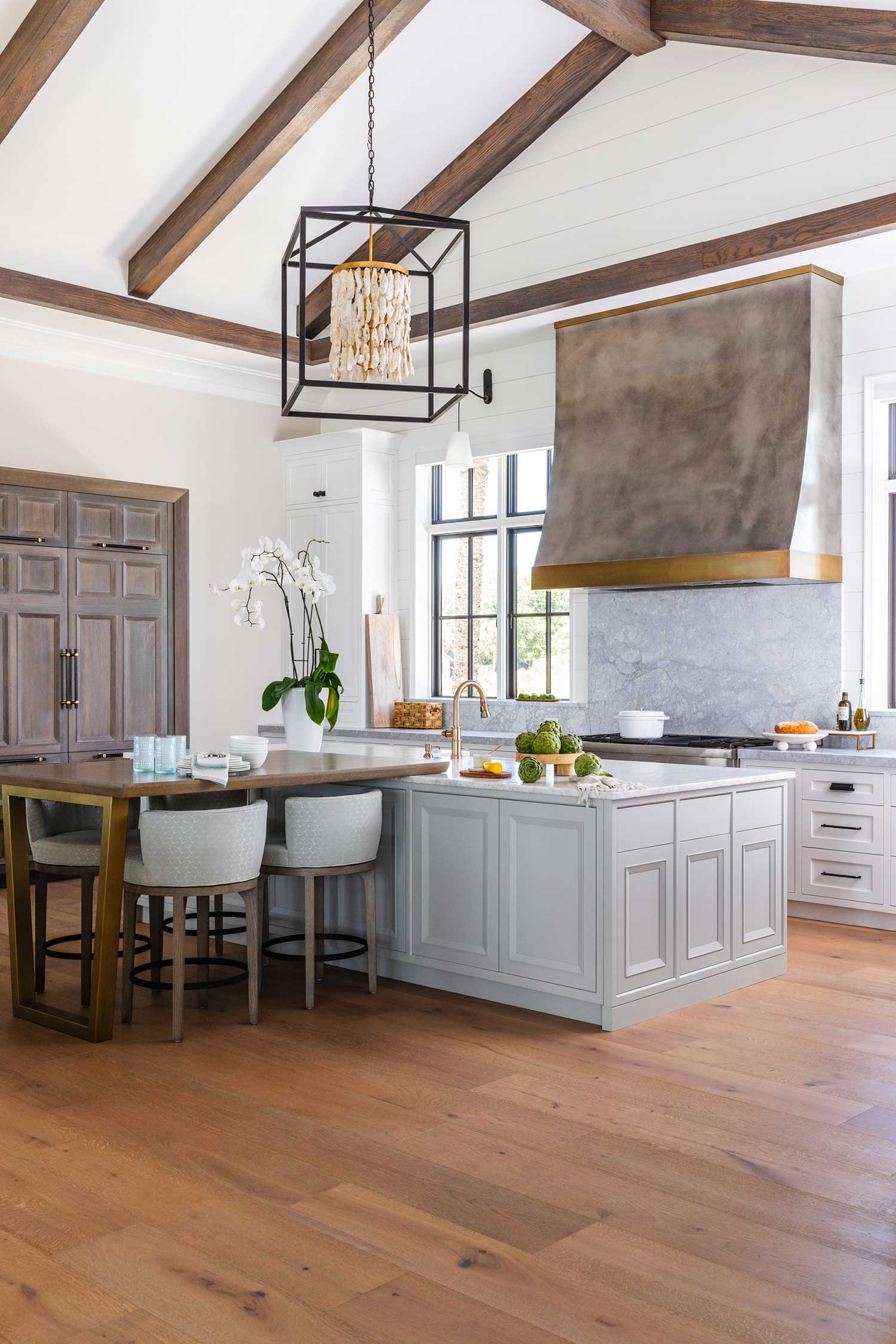 A modern kitchen with exposed wood beams, a large island with gray cabinets, beige upholstered stools, a wooden floor, stainless steel appliances, a large vent hood, and an orchid on the counter. Natural light fills the room.