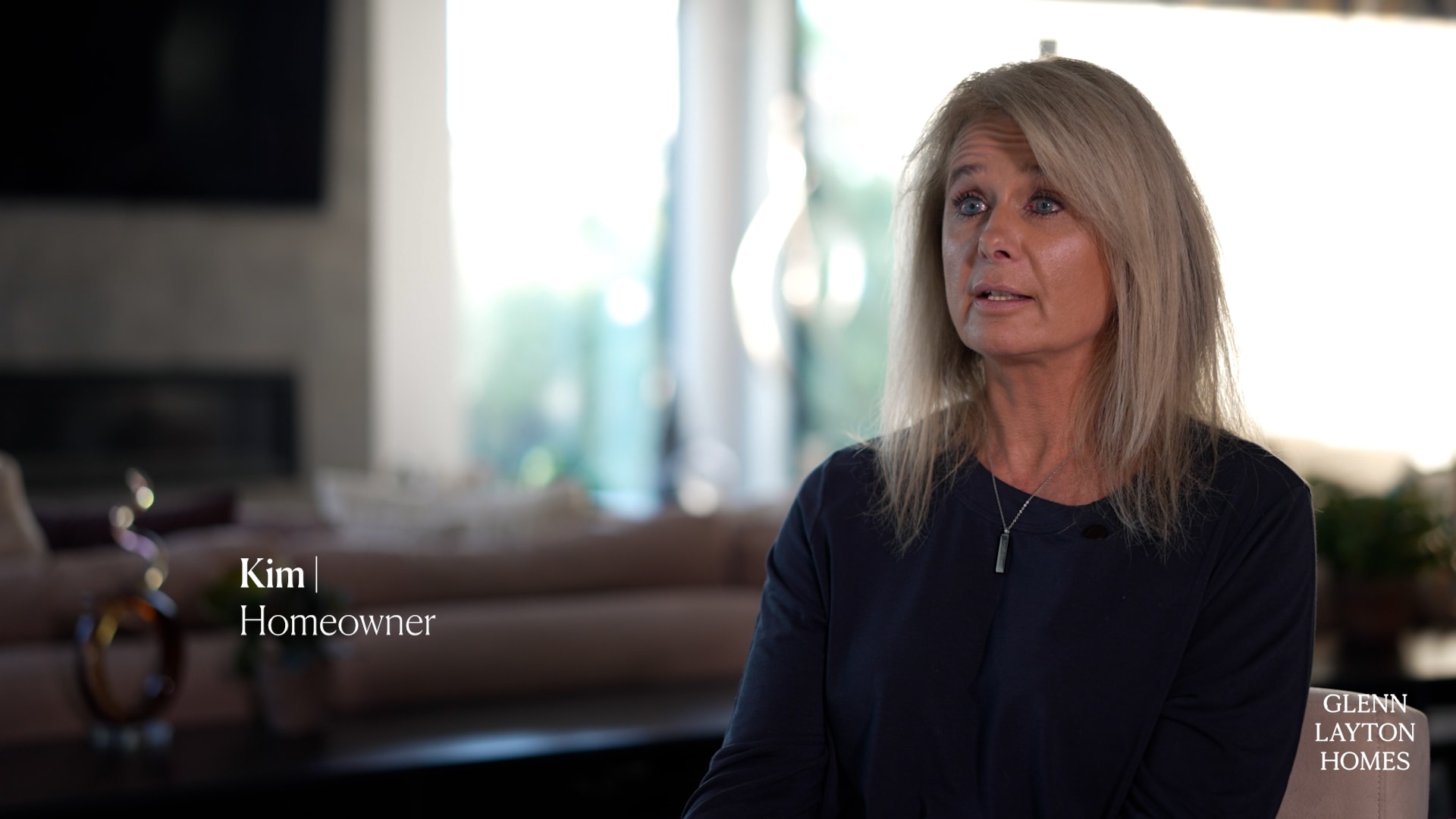 A woman with long blonde hair, wearing a dark top, sits indoors and looks slightly upward. Text on the image reads Kim | Homeowner. The Glenn Layton Homes logo, a custom home builder in Florida, appears in the lower right corner.