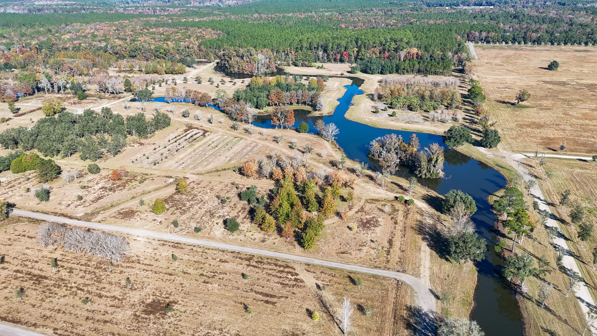 Aerial view of a rural landscape with fields, trees, winding dirt roads, and small ponds under a clear sky. The scene shows autumn colors and a mix of open land and wooded areas.