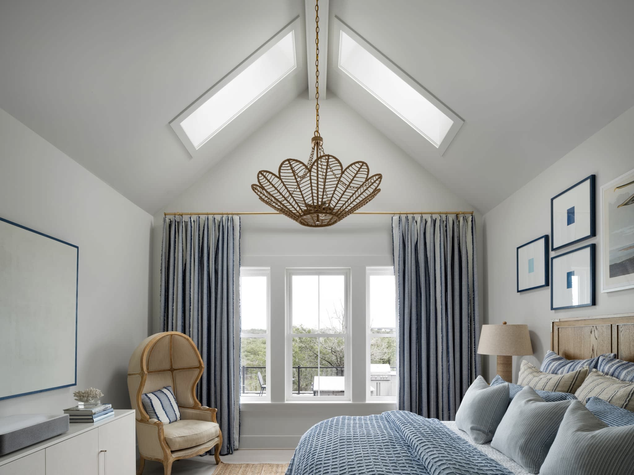 A bright bedroom with vaulted ceiling, two skylights, a large rattan chandelier, blue-and-white bedding, striped curtains, a light wood chair, and framed art on the walls. Large windows overlook greenery outside.