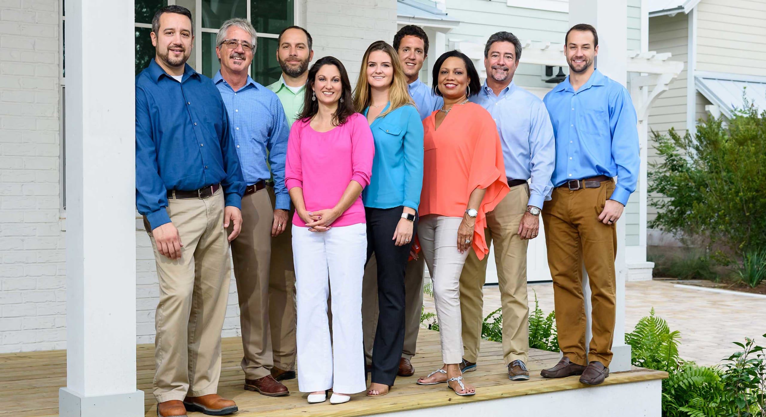 A group of nine adults, five men and four women, stand together on a porch in front of a house. They are smiling and dressed in business casual attire in various shades of blue, pink, and beige.
