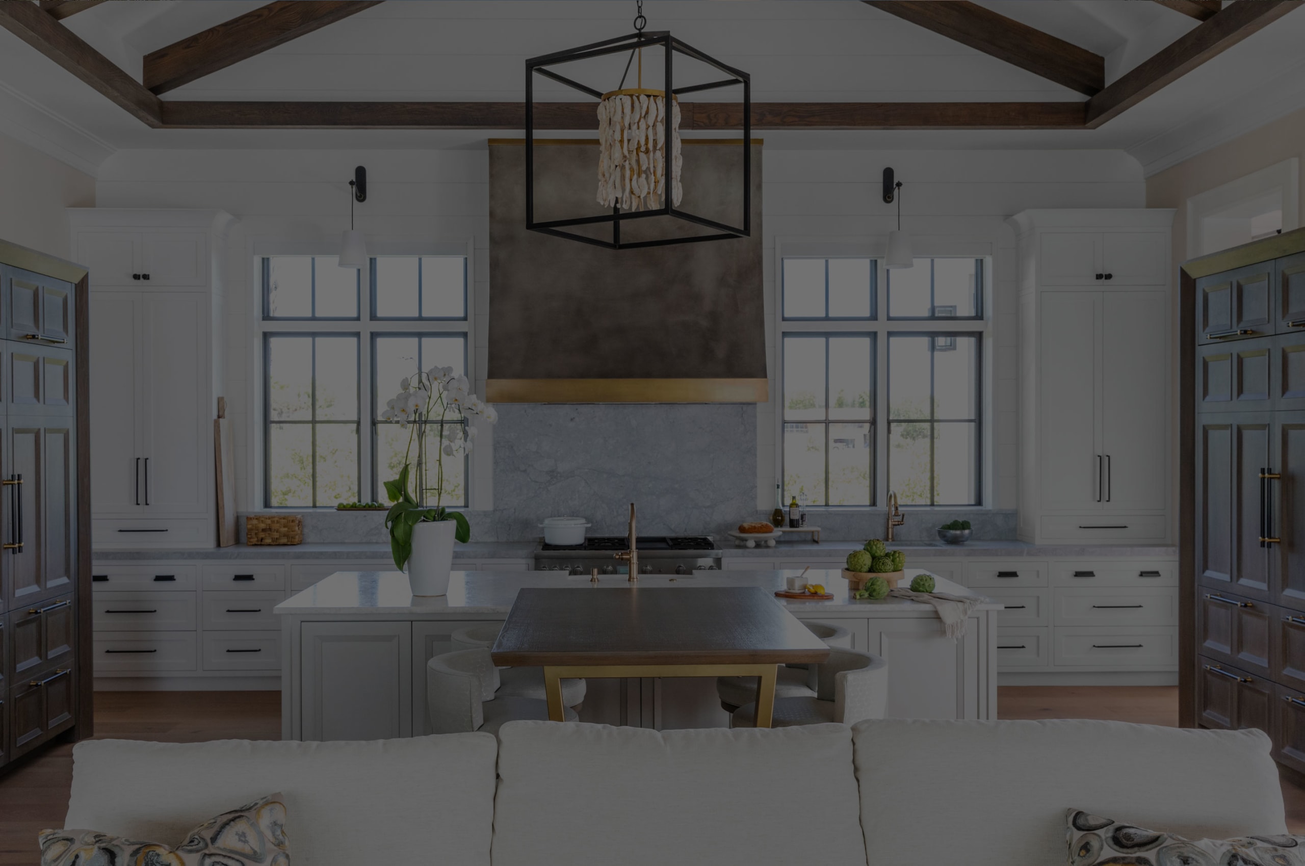 Modern kitchen by a custom home builder in Florida features exposed wooden ceiling beams, white cabinetry, a central island with plants, marble countertops, large windows for natural light, and a geometric pendant light above the island.