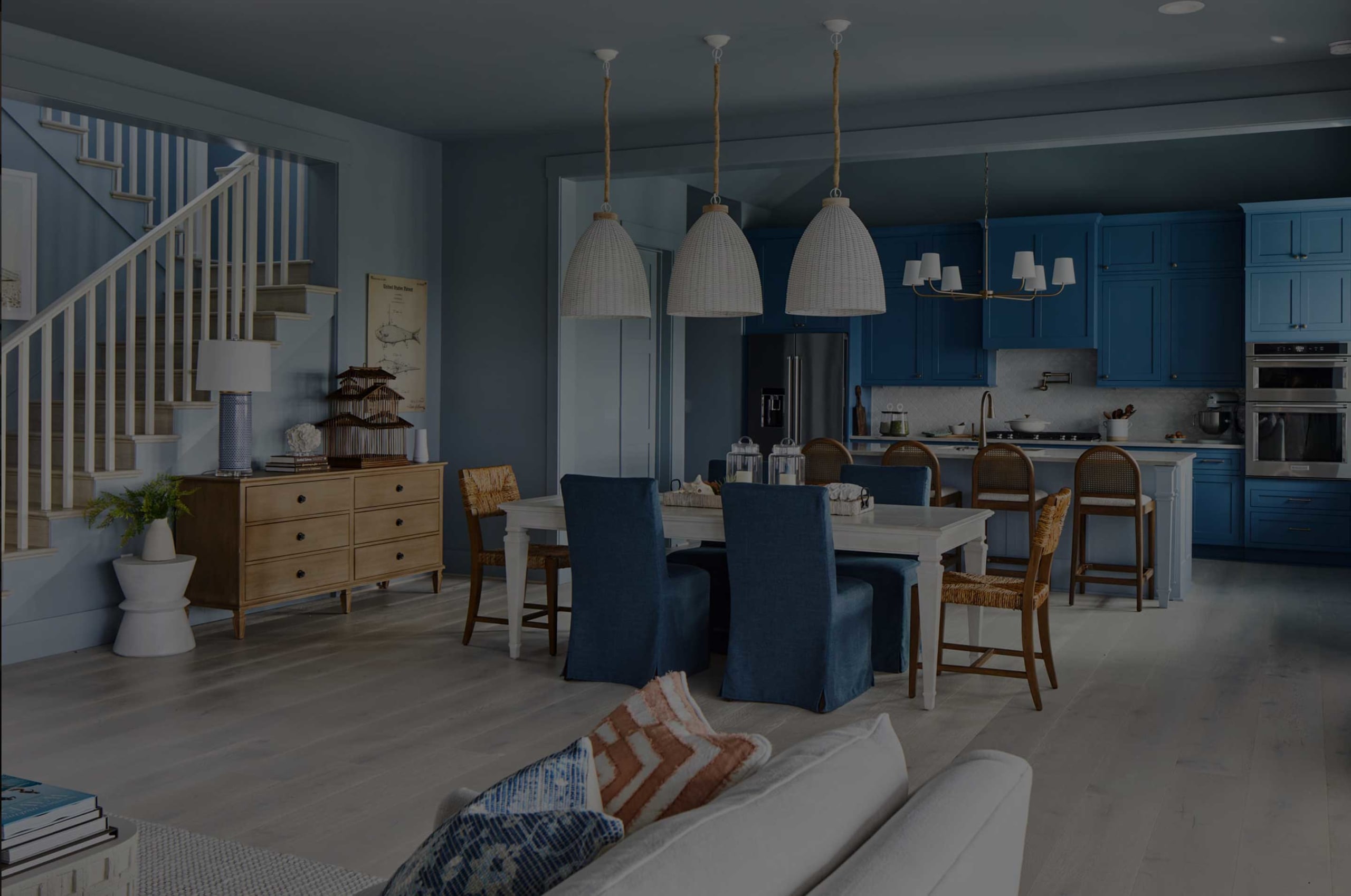 A modern open-concept kitchen and dining area by a custom home builder in Florida, featuring blue cabinets, a white island, woven pendant lights, wooden chairs, a staircase, and a mid-century sideboard with decor.