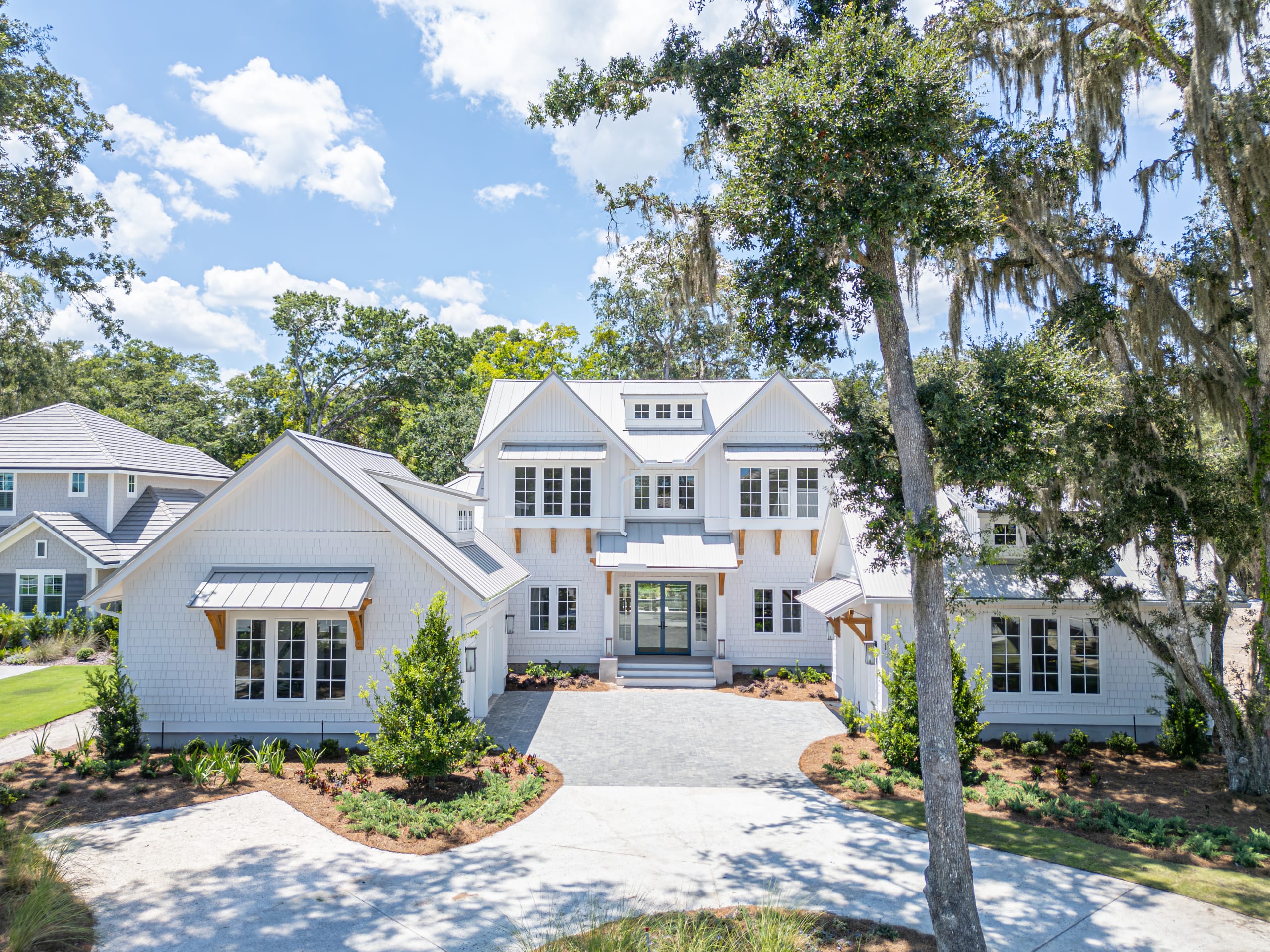 A large, modern white house with multiple dormer windows and a gray shingle roof, surrounded by trees and landscaping, under a blue sky with scattered clouds—an ideal example of homes in Nocatee FL.