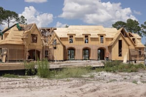 A large house under construction, with wooden framing and plywood sheathing exposed. Construction materials and equipment are visible, and trees stand in the background under a partly cloudy sky.