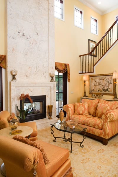 Elegant living room with high ceiling, tall marble fireplace, large windows, ornate orange sofas, glass coffee table, stairway with dark railing, decorative lamps, and framed art on pale yellow walls.