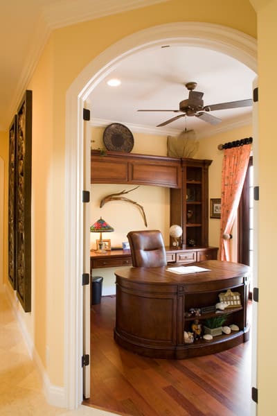 A home office with yellow walls, a wooden desk and chair, built-in shelves, and a ceiling fan. The desk holds a lamp, globe, and framed photos. Light enters through a window with coral curtains.