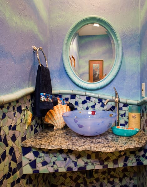 Colorful bathroom with mosaic tile walls, a granite countertop, a frosted glass vessel sink, a shell decoration, a blue towel on a ring, and a round mirror reflecting framed art and ceiling lights.