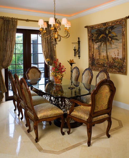 Elegant dining room with a glass-top table, eight ornate wooden chairs, a chandelier, gold curtains, French doors, and a wall tapestry depicting a tropical beach scene. A vase with flowers is centered on the table.
