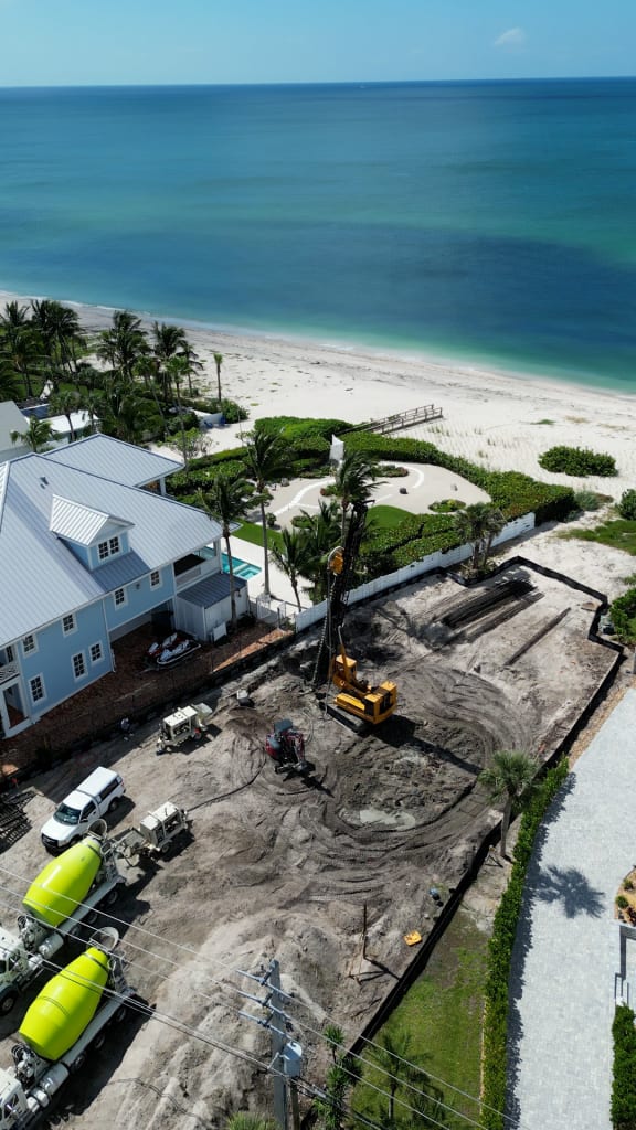 Aerial view of a beachfront construction site with heavy machinery and trucks near a white house, bordered by palm trees and a sandy beach with calm blue ocean water in the background.