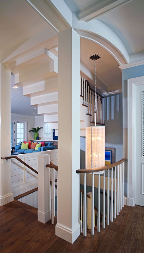 Modern interior with a wooden staircase, white railings, and a hanging cylindrical light fixture. In the background, a living area with colorful cushions on the sofa and bright natural light.