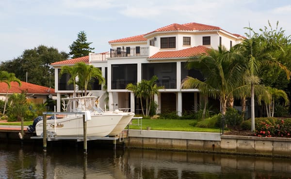 A large, white, three-story house with red-tiled roof sits beside a canal, surrounded by palm trees and greenery. A white boat is docked at a private pier in front of the house.