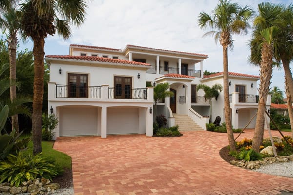 A large, white, two-story house with terracotta roof tiles, double garages, balconies, and a wide brick driveway, surrounded by palm trees and lush landscaping under a partly cloudy sky.