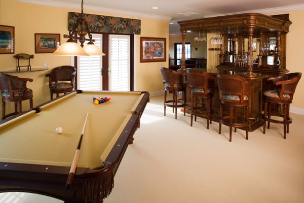 A home recreation room with a billiards table set for a game and a wooden bar area with several high chairs. The room is brightly lit, with framed art on yellow walls and large windows with white shutters.