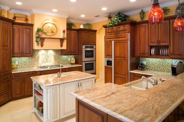Spacious kitchen with wooden cabinets, granite countertops, a white island, built-in double ovens, a stovetop with decorative backsplash, and hanging red pendant lights. Plants and decorative items are displayed above the cabinets.