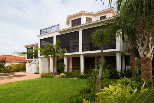 A two-story house with a screened-in porch, white exterior, and red tile roof, surrounded by green grass, tropical plants, palm trees, and a brick patio area.