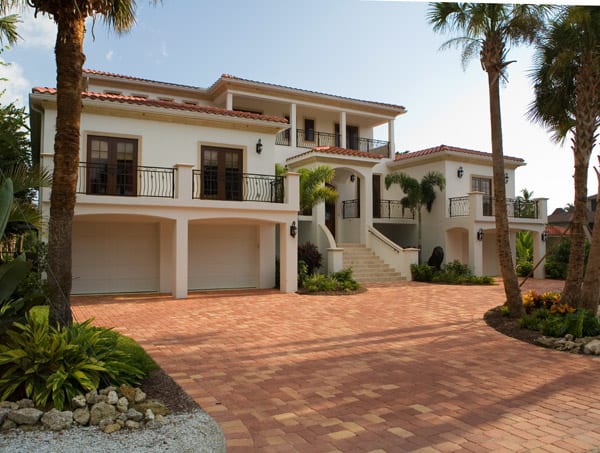 A large, modern Mediterranean-style house with white walls, red-tiled roof, balconies, and a double garage. The driveway is paved with red bricks, and the property is surrounded by lush greenery and palm trees.
