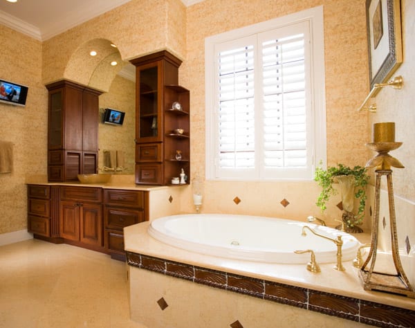 Luxurious bathroom with a large oval bathtub, gold fixtures, wooden cabinets, a vanity with sink, decorative plants, candles, and two wall-mounted TVs. Sunlight streams in through white shuttered windows.