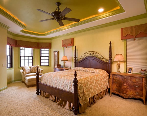 Elegant bedroom with a wooden bed frame, floral bedding, ornate nightstands with lamps, armchair by large windows, beige carpet, tray ceiling with recessed lighting, and ceiling fan.