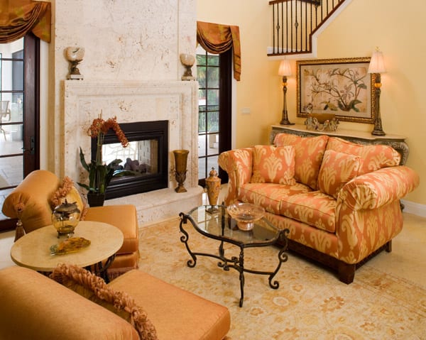 Elegant living room with orange patterned sofa and chairs, a glass coffee table with a metal base, a marble fireplace, and decorative accents, all in warm tones with natural light from large windows.