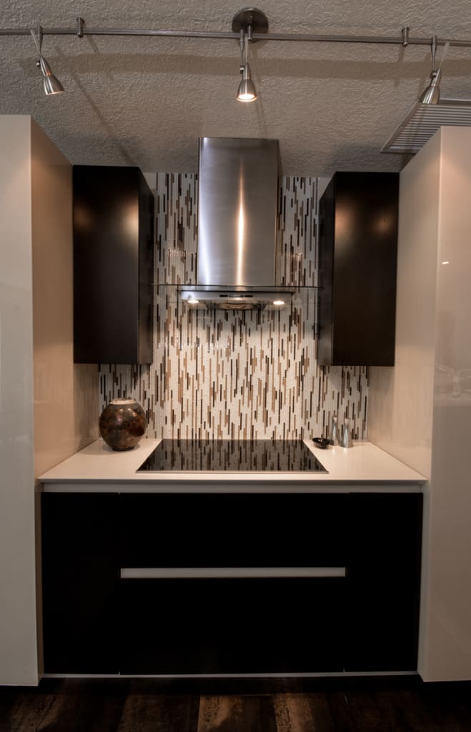 Modern kitchen with a sleek black electric cooktop, stainless steel hood, dark cabinets, and a decorative vertical tile backsplash. A round decorative object and utensils sit on the white countertop.