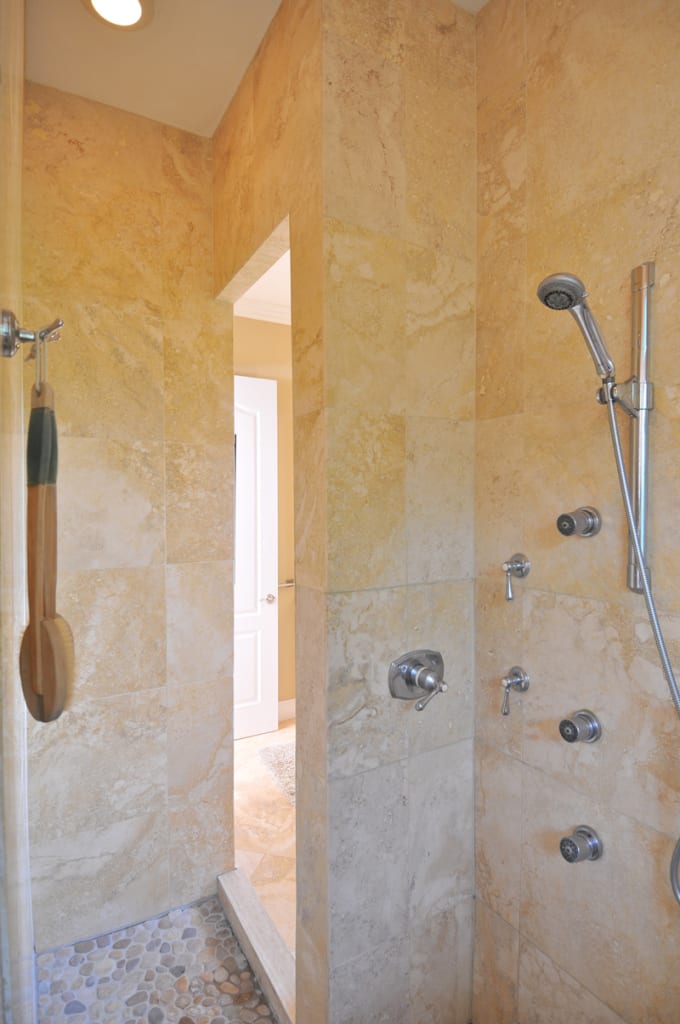 A walk-in shower with beige stone tile walls, multiple chrome shower controls, a handheld showerhead, built-in body sprays, and a wooden bath brush hanging on a hook. Pebble-style flooring covers the shower floor.