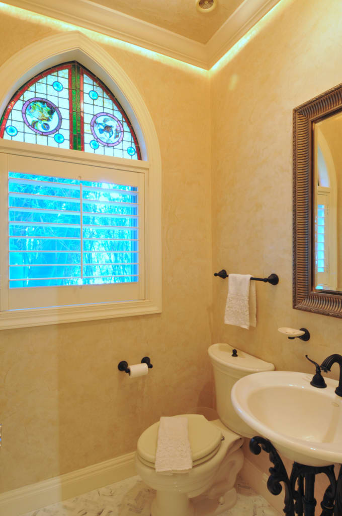 Elegant bathroom with beige walls, a toilet, towel rack, ornate mirror, and a round sink on a black stand. A large window with colorful stained glass and white shutters lets in natural light.