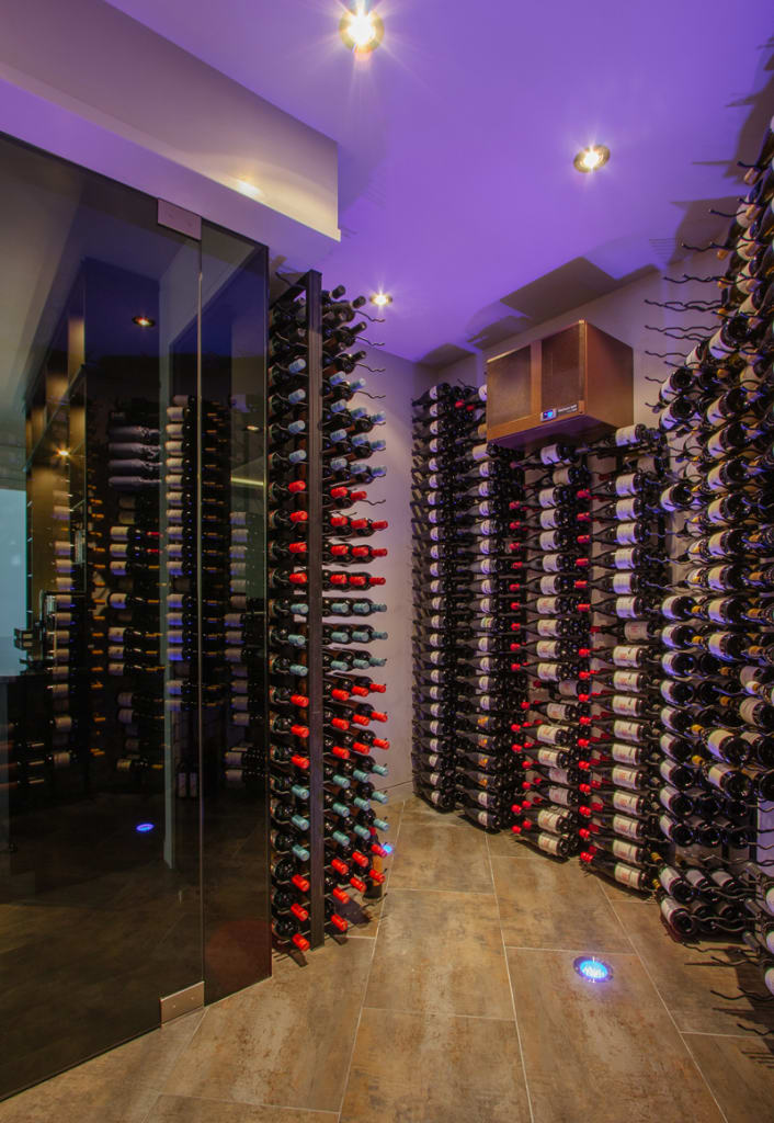 A modern wine cellar with floor-to-ceiling racks holding many wine bottles, glass door entrance, warm lighting, and wood-look tile flooring. The room is illuminated with purple-toned lights.