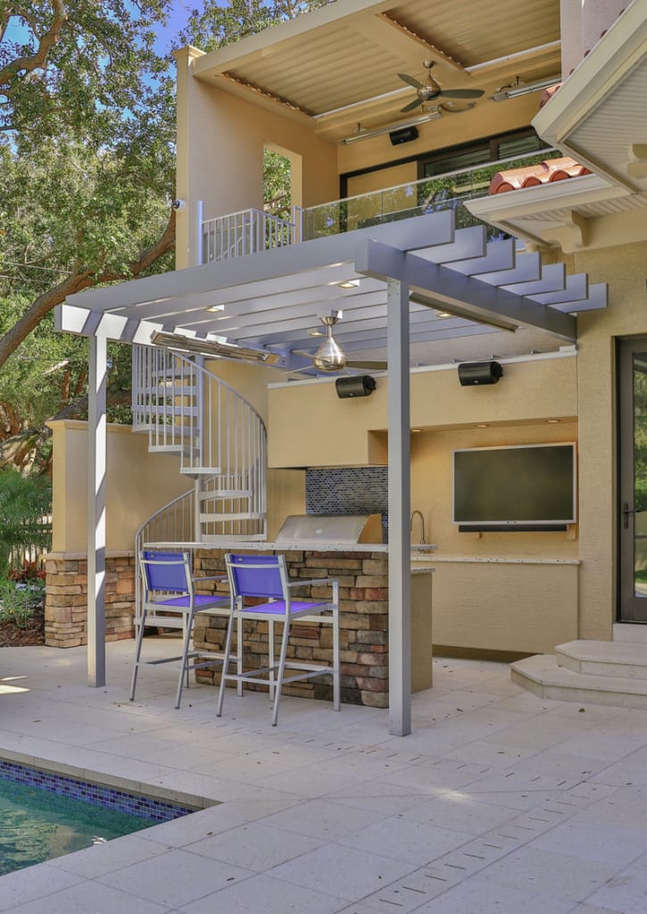 Outdoor poolside kitchen with a pergola, spiral staircase, two barstools, grill, flat-screen TV, and upper balcony with ceiling fans, surrounded by trees and modern architecture.